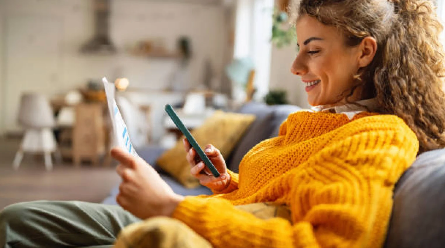 Woman looking at internet bill on her phone and on paper and smiling in a yellow sweater.