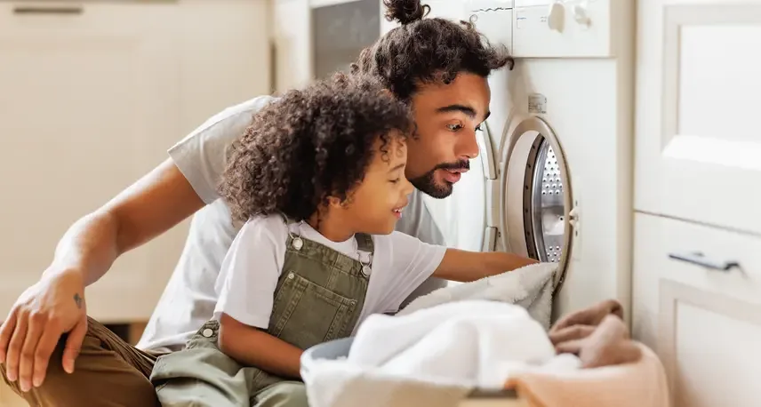 A man and a child sitting down next to each other while putting towels into a dryer