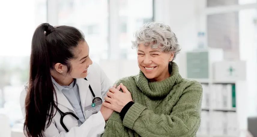 A woman with brown hair in a pony tail wearing a lab coat and stethoscope is smiling and looking at another woman with gray hair in a green sweater while touching hands