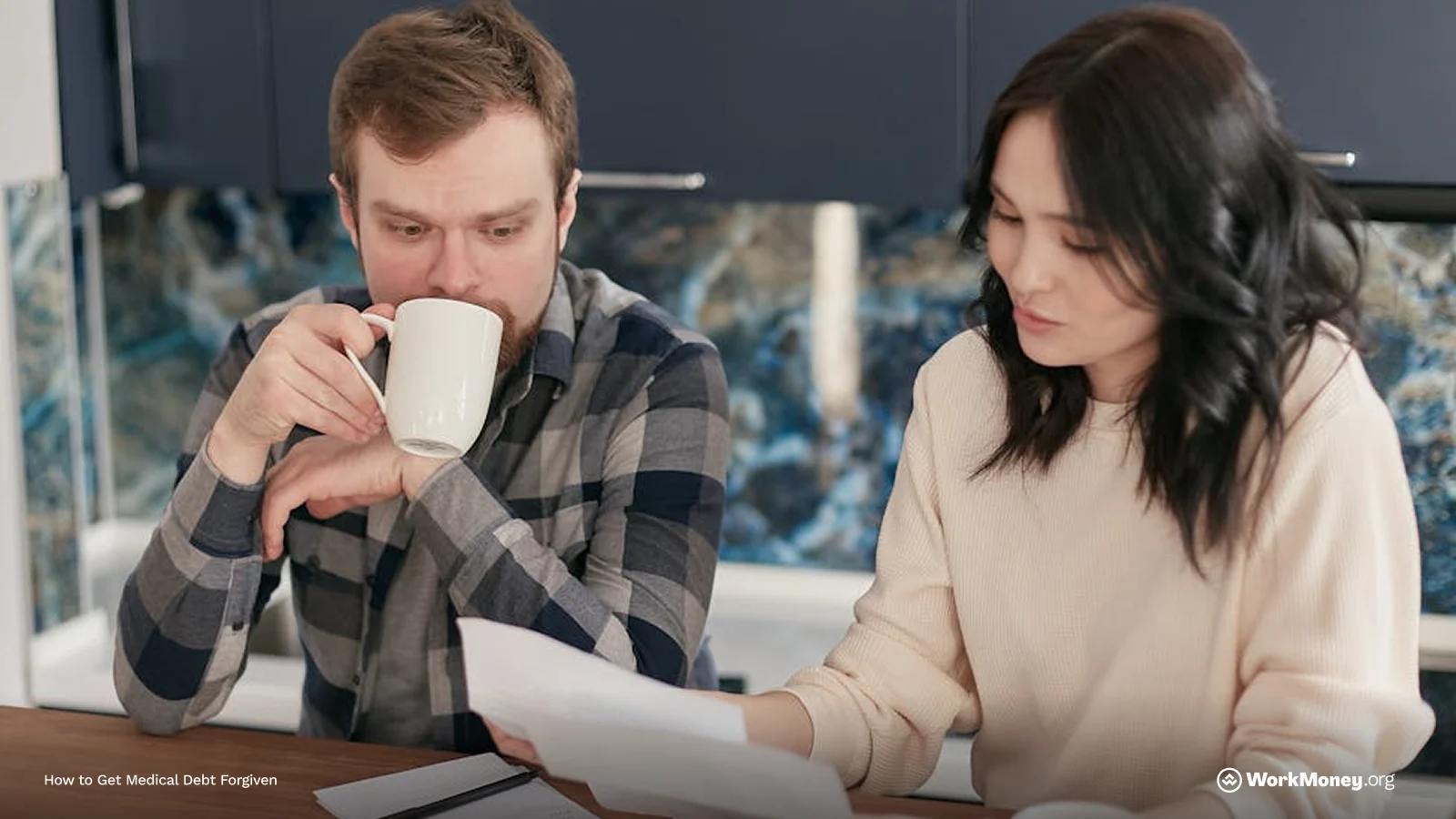 A woman reads a document while a man beside her drinks from a mug and looks worried, both appearing focused and tense at the kitchen table.
