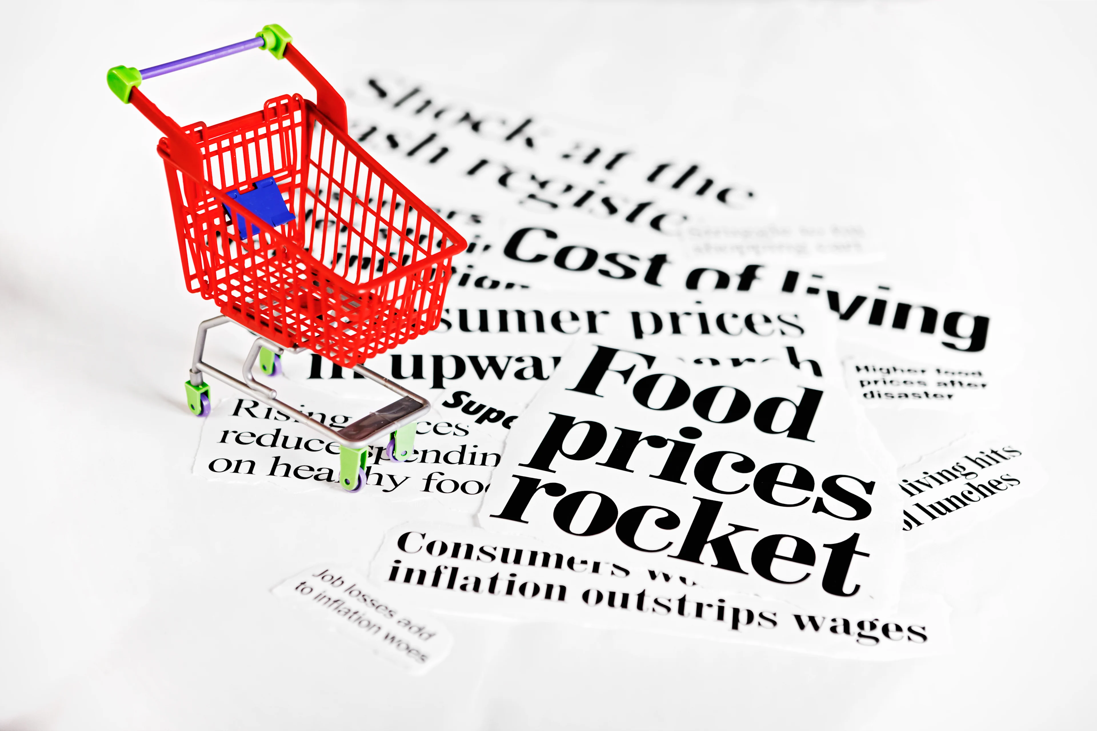 A small red shopping cart sits on a white surface surrounded by newspaper clippings with bold headlines about rising living costs and inflation. Visible phrases include “Food prices rocket,” “Cost of living,” and “Consumer prices up.” The image symbolizes inflation and the increasing cost of everyday goods.