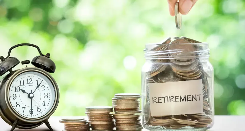 An alarm clock next to a pile of coins and a jar with more coins in it. The jar has a label that says Retirement and there is a hand putting another coin into the jar.