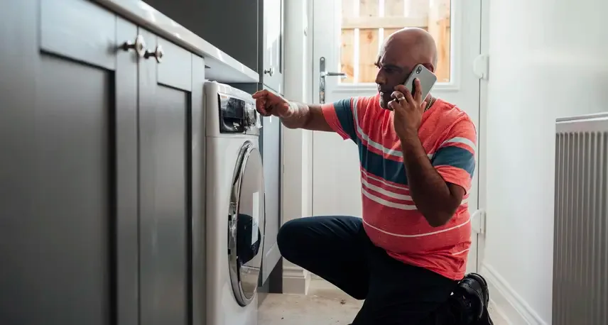 A man with a red striped shirt kneeling down and touching a washing machine while holding a cell phone to his ear