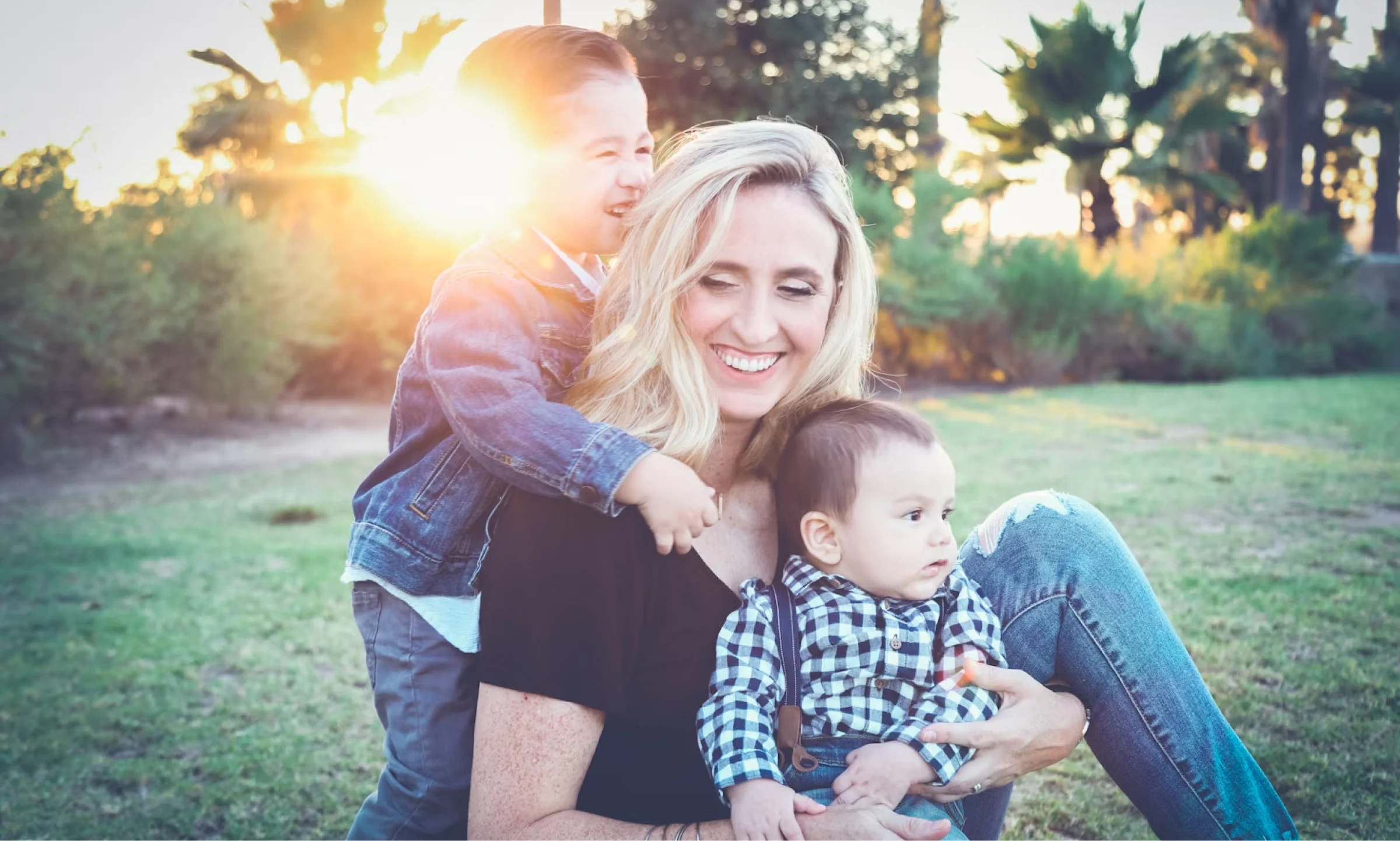 A woman with blond hair and a black tshirt smiling while sitting outside. She is holding a baby and another child is hugging her from behind while smiling.