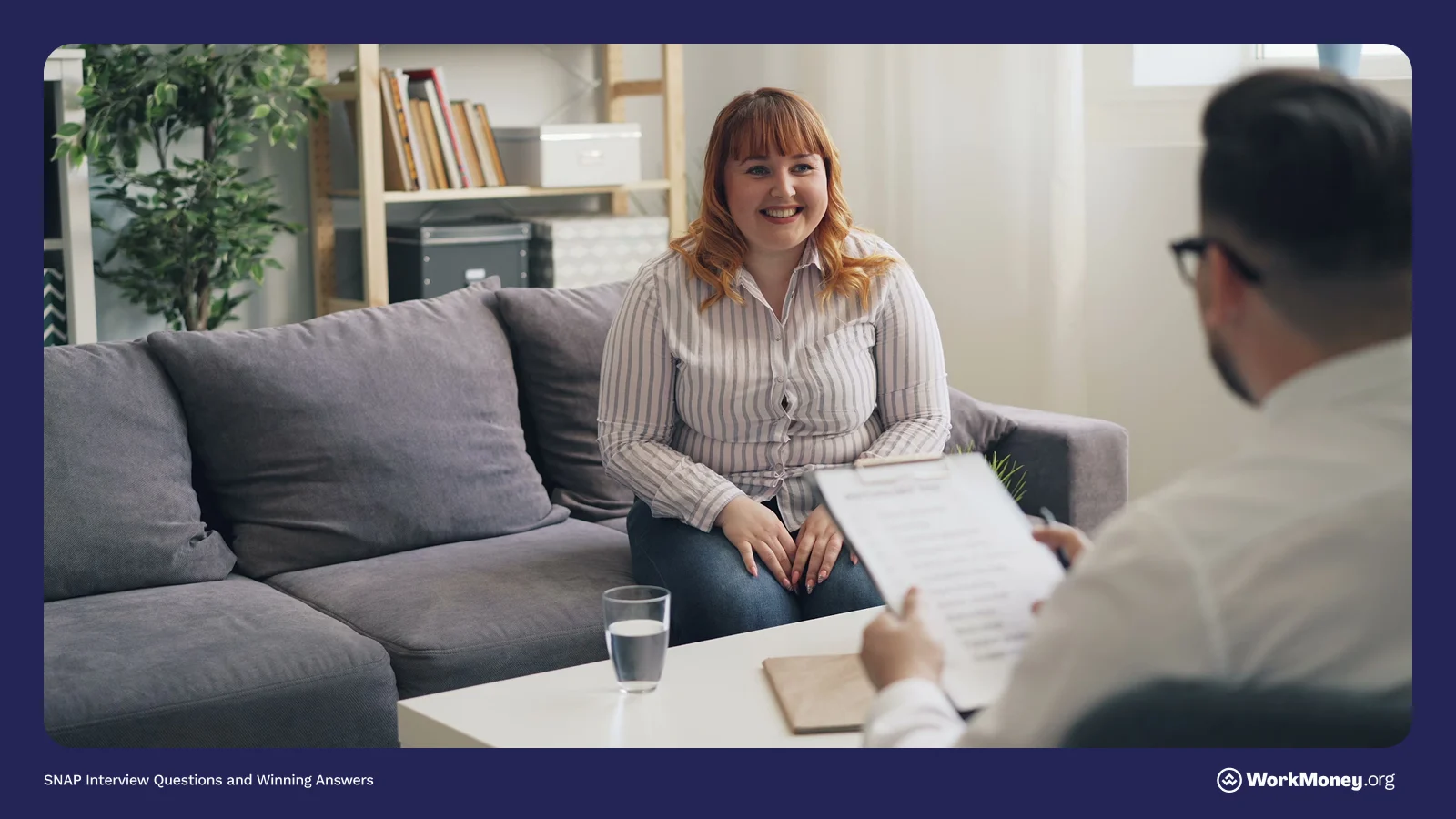 A woman sits on a couch smiling during an interview, facing a man who is holding a clipboard with a printed form. The setting appears to be a casual office or consultation room, with bookshelves and a plant in the background.