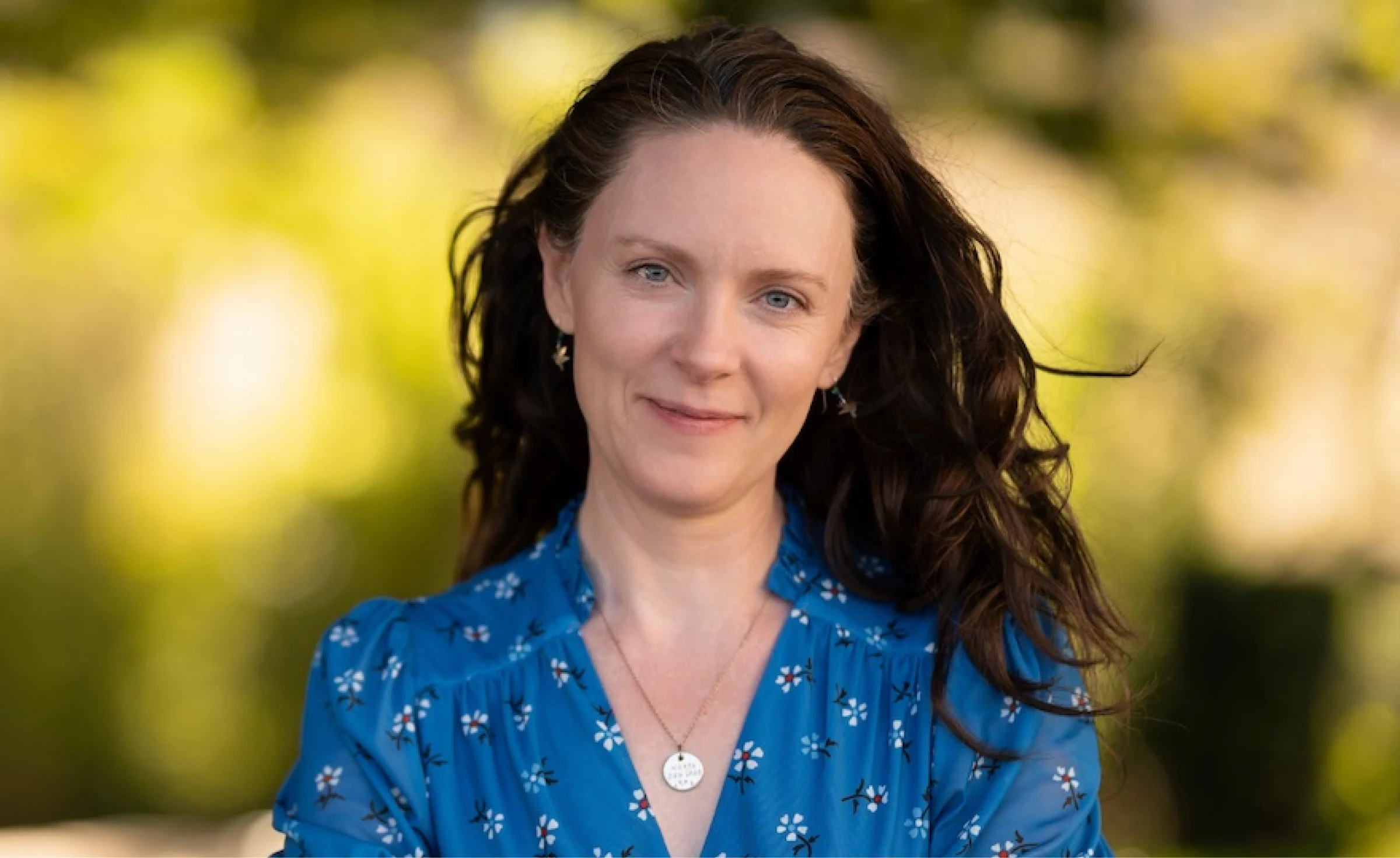 A woman with dark hair and a blue blouse smiling slightly outdoors