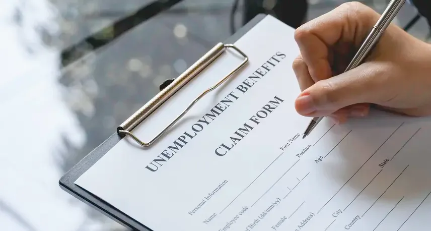 A hand with a pen writing on a form on a clipboard that says Unemployment benefits claim form