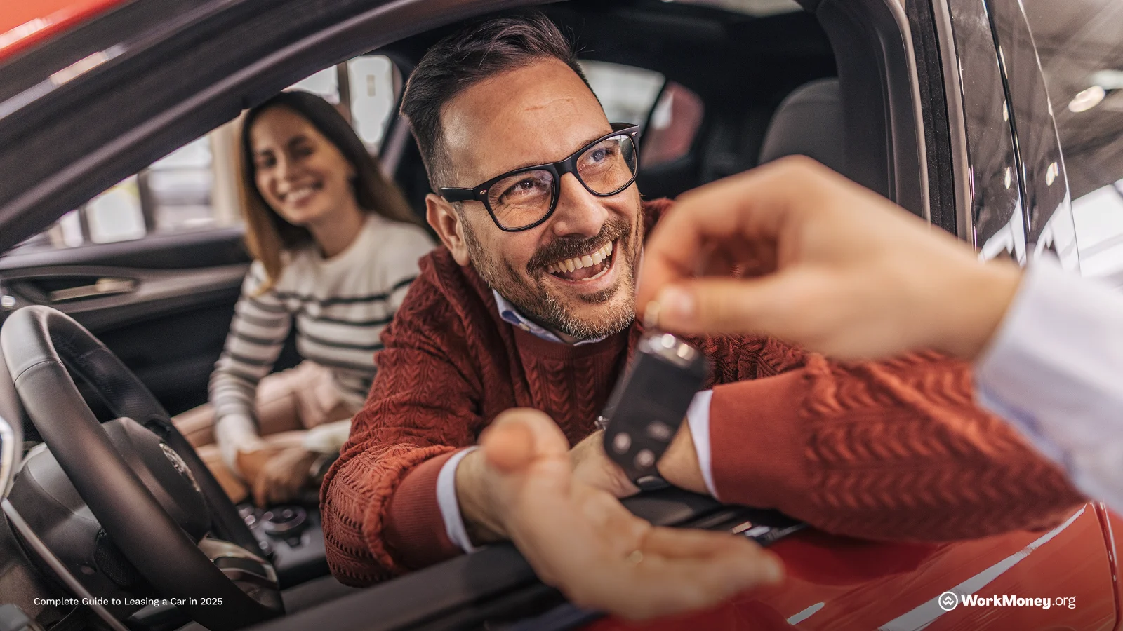 Man in orange sweater is handed keys while sitting in the driver's seat