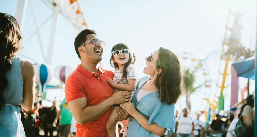 2 people holding a child smiling with sunglasses on in front of a rollercoaster