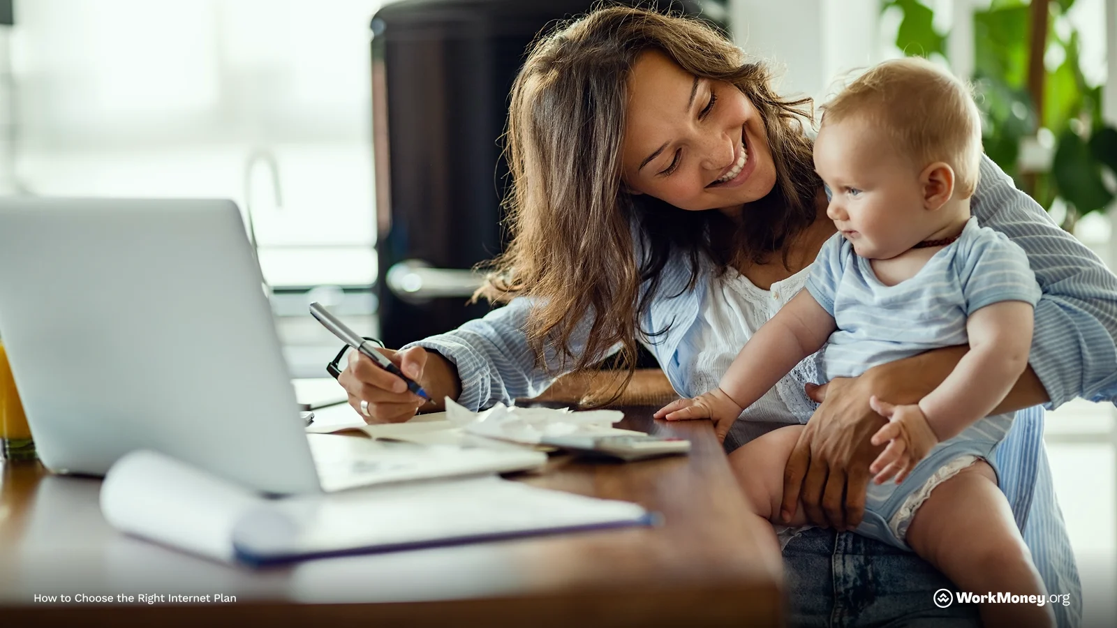 A smiling woman sits at a table working on a laptop while holding a baby on her lap. She’s writing on paper with a pen, surrounded by documents and a cup of juice. The setting appears to be a bright home kitchen. Text in corner reads “How to Choose the Right Internet Plan.”