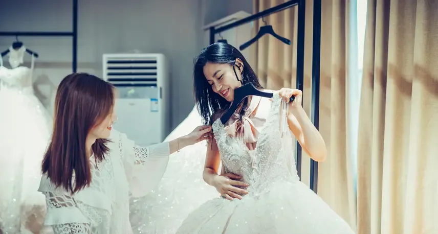 A woman with dark hair holding up a white dress on a hanger. Another woman is looking at the dress and smiling.