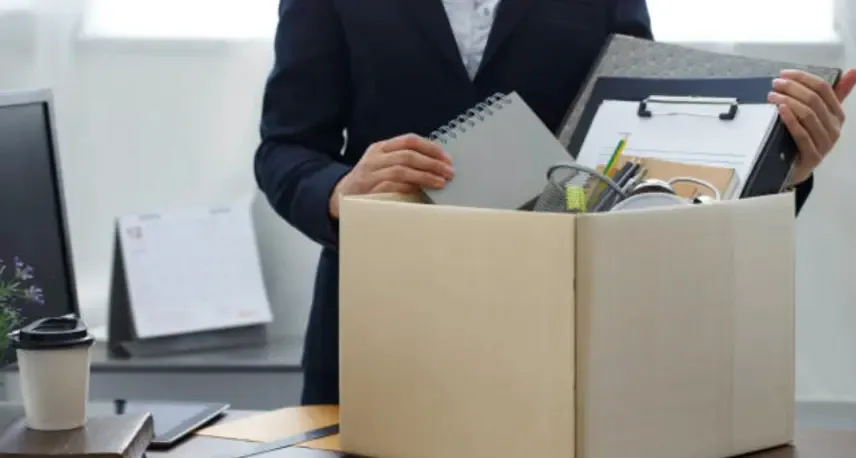 A person in a dark suit holding various items in a cardboard box on a desk