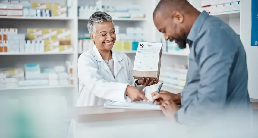 A person in a white lab coat pointing at a pad of paper. Behind the person is a shelf with various boxes. In front of her is a person smiling while writing on the paper she's pointing at.