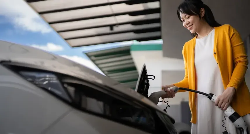 A woman with dark hair and a yellow sweater smiling while holding a charger attached to a gray car