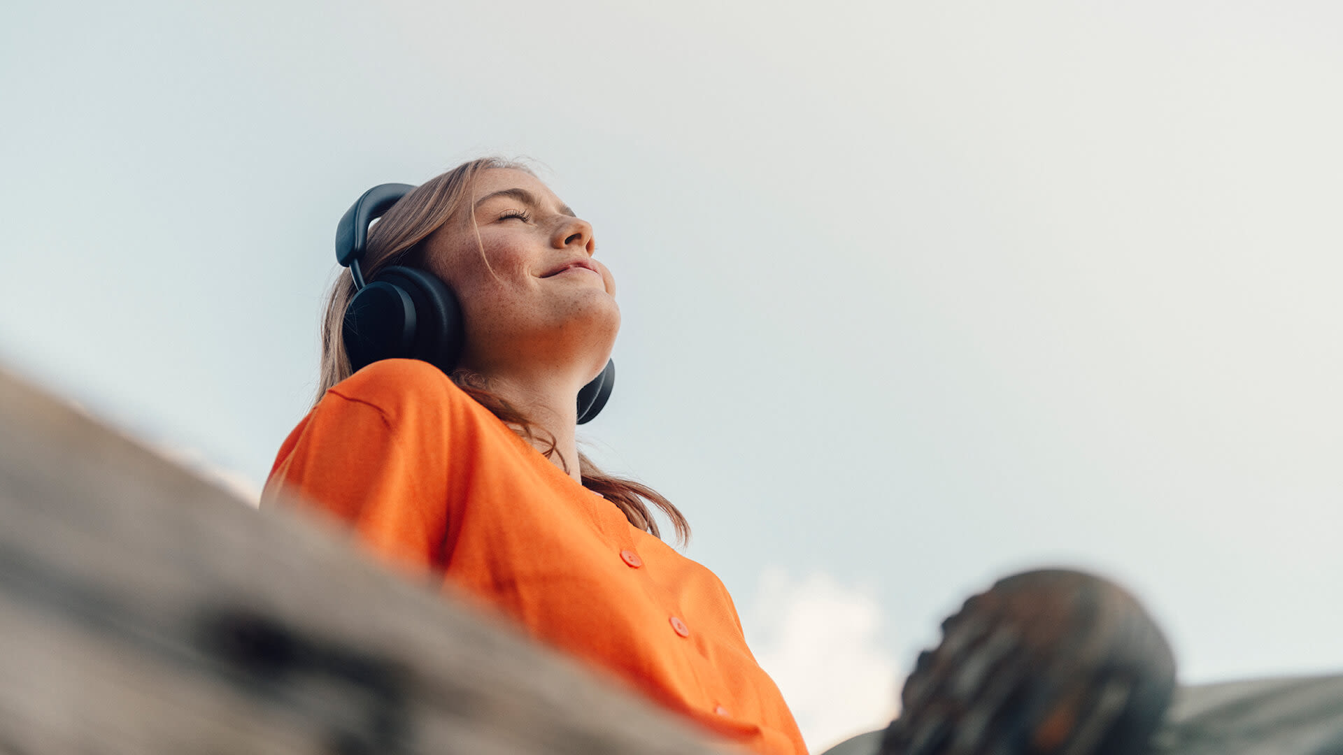 Kvinna i orange tröja sitter utomhus och lyssnar på musik i ett par stora hörlurar. Hon blundar och ler.