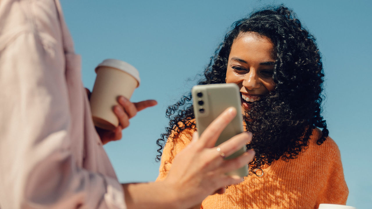 Person håller i kaffemugg och mobil medan en tjej med lockigt hår och orange tröja tittar på mobilen när hon skrattar.
