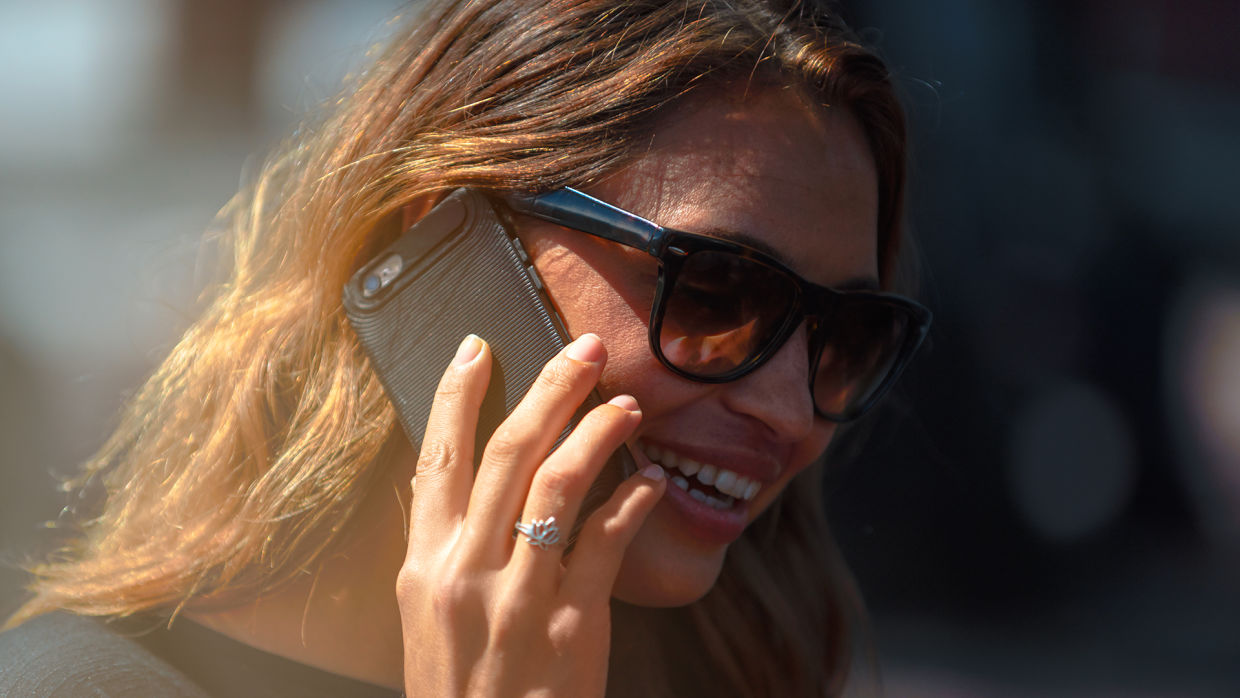 Kvinna i brunrött hår och solglasögon som pratar i mobilen och ler.