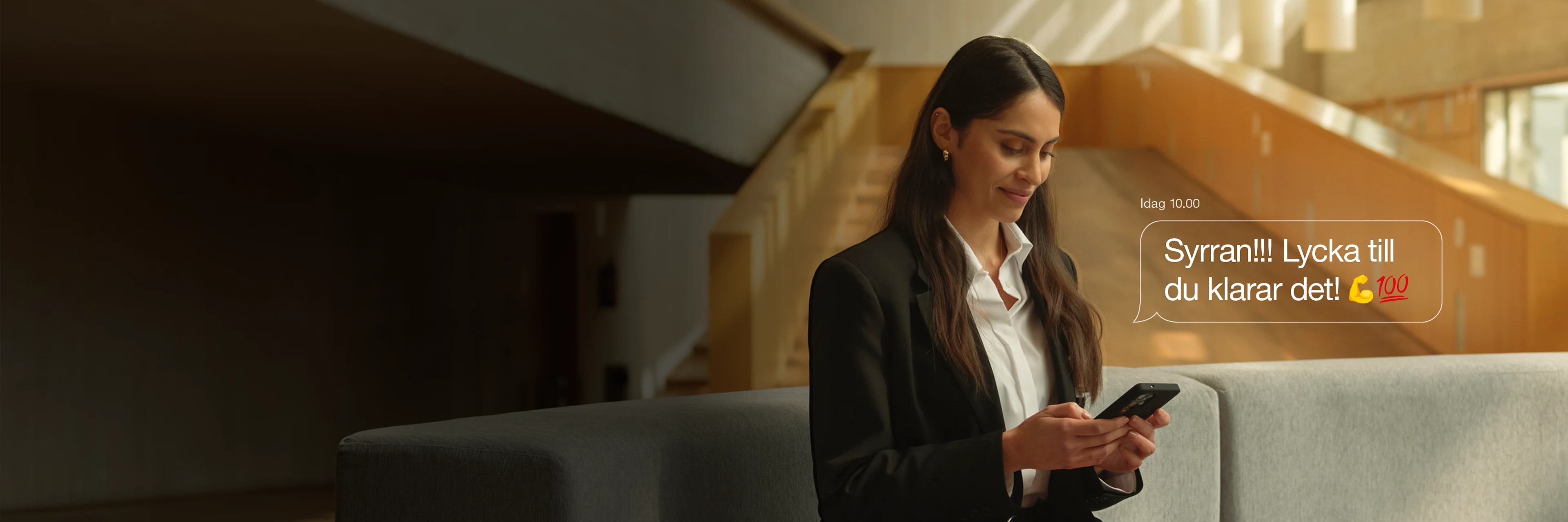 Kvinna i kostym sitter på soffa i ljust rum, ler och tittar på sin mobil, med peppande sms på skärmen.