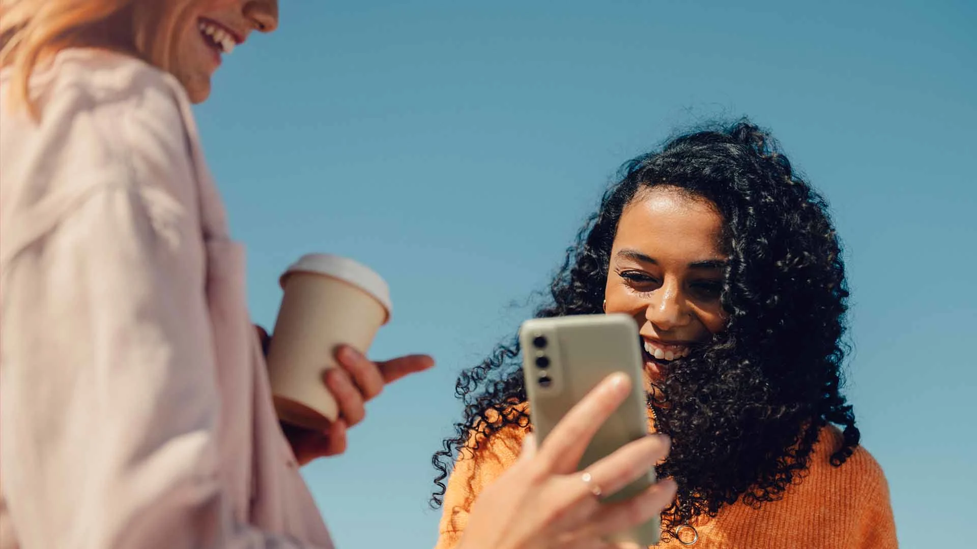 Två kvinnor utomhus skrattar tillsammans, en håller upp en mobil mot den andra som leende tittar in i den. Hon som håller upp mobilen har en take away kaffekopp i andra handen. Bakgrunden är en blå himmel.