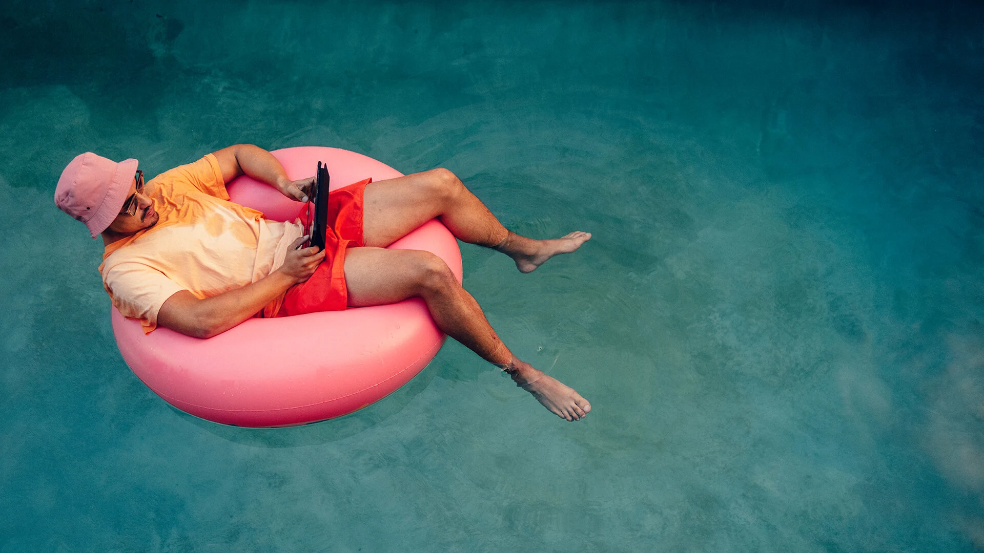 Man surfar på sin surfplatta i poolen. Han ligger på en rosa badring med fötterna i det turkosa vattnet.