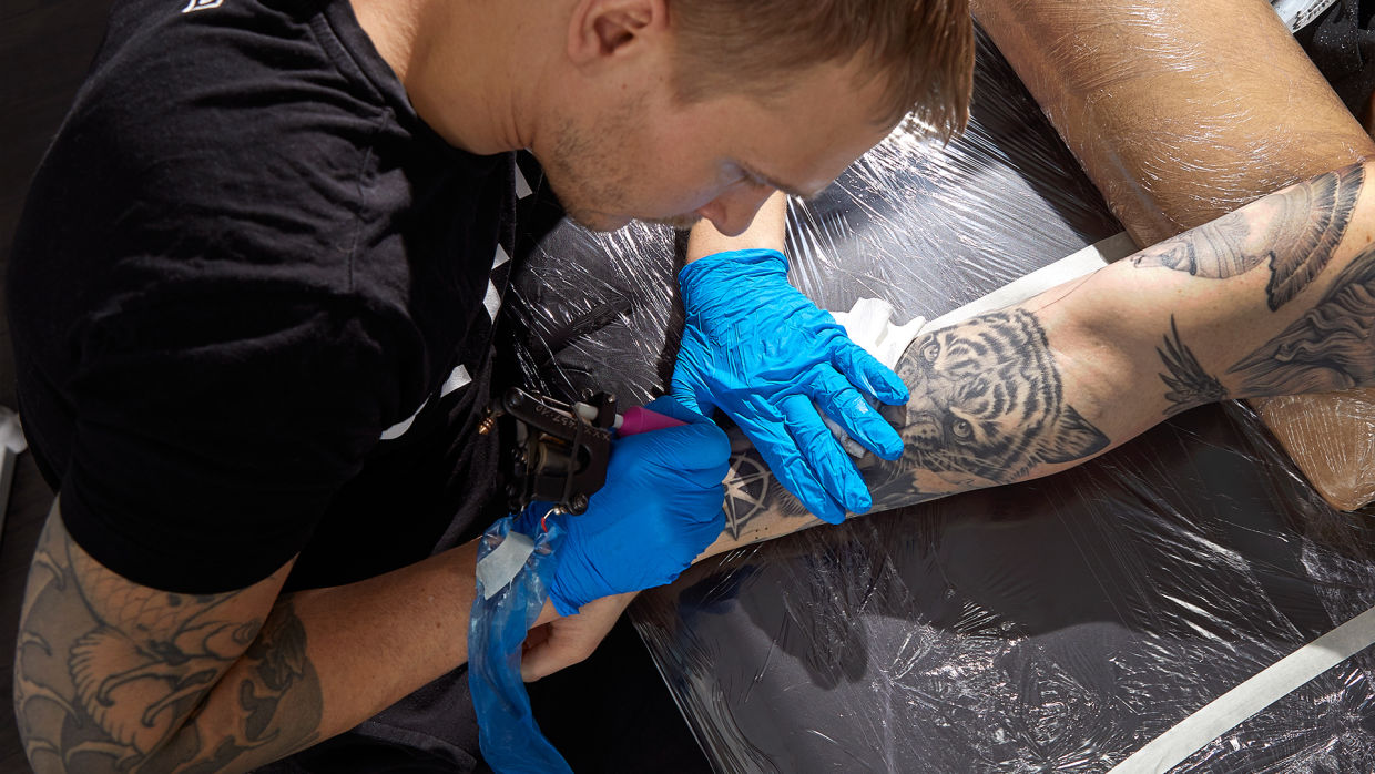 Tatuerare i svart t-shirt och blå handskar i nitril tatuerar en underarm på en svart brits.
