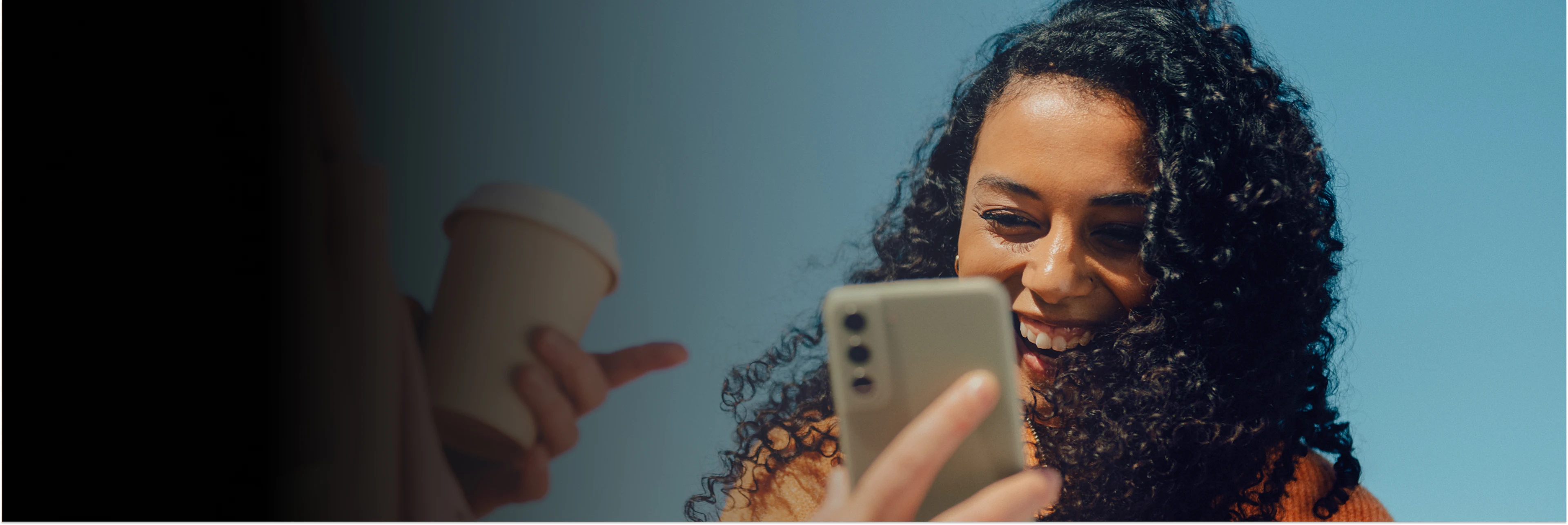 Leende kvinna med lockigt hår tittar på en mobil som hålls upp av en annan person, utomhus. Personen som håller upp mobilen har en take away kaffekopp i andra handen. Bakgrunden är en blå himmel.