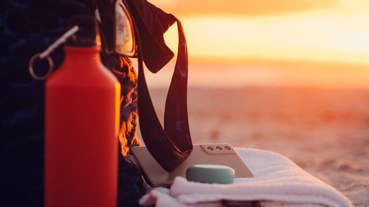 En knallröd vattenflaska står på stranden i solnedgången tillsammans med en träningsväska, mobil och ett par hörlurar.