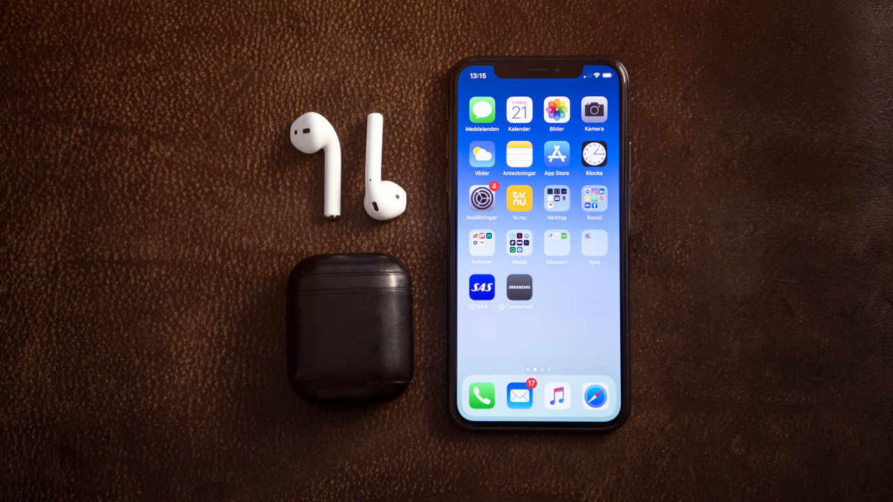 En mobiltelefon av modellen Apple iPhone, ett AirPods-fodral samt vita AirPods ligger på en brun matta i skinn.