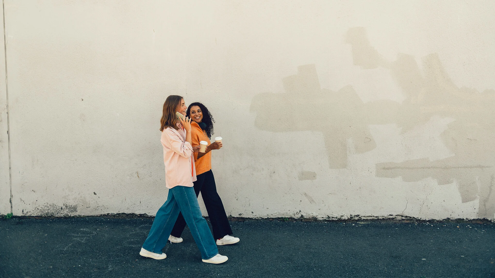 Två tjejer går med varsin kaffe i handen. Den ena pratar i telefon. 