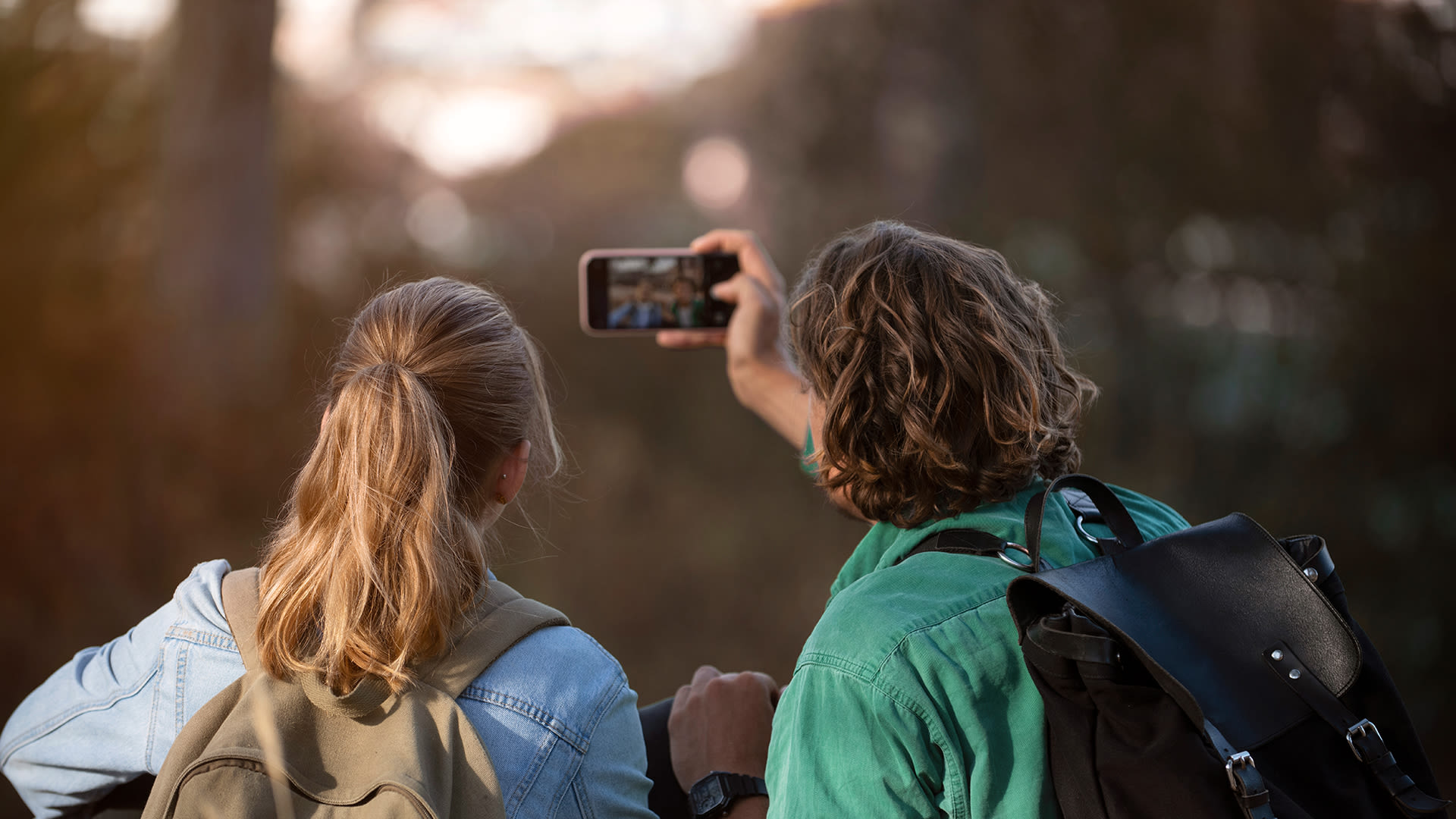 En tjej med långt hår och en kille i grön skjorta är ute i skogen. Han håller upp sin mobiltelefon för att ta ett kort. 