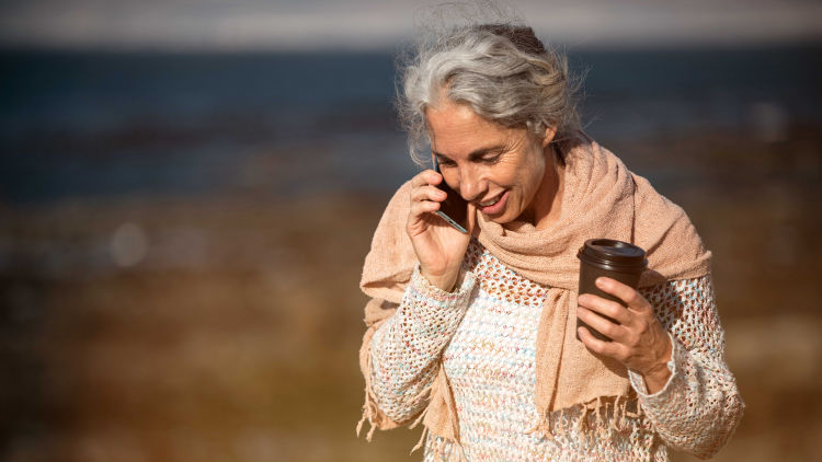 En kvinna med grått, yvigt hår går ute i naturen och pratar i telefon och dricker kaffe samtidigt. Hon har en beige sjal virad runt halsen.