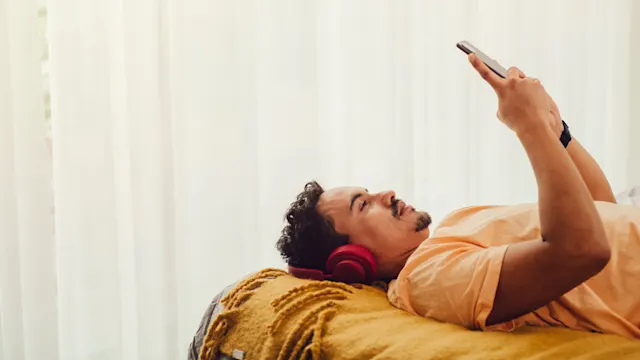 En man med hörlurar ligger på en säng och tittar på en mobil.