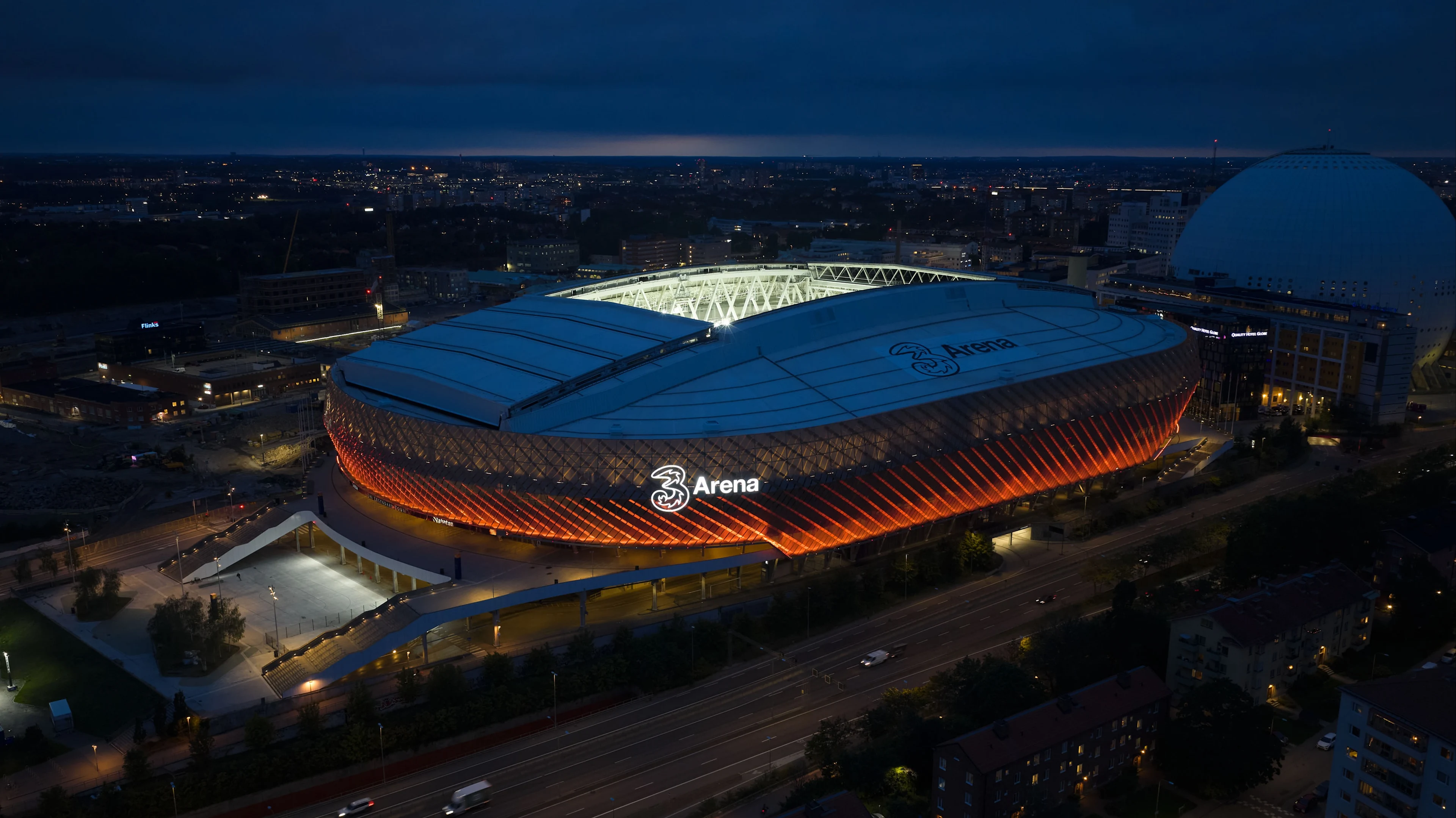 Flygfoto av en upplyst arena i skymningen, med orange och vita ljus, omgiven av stadens byggnader och vägar.