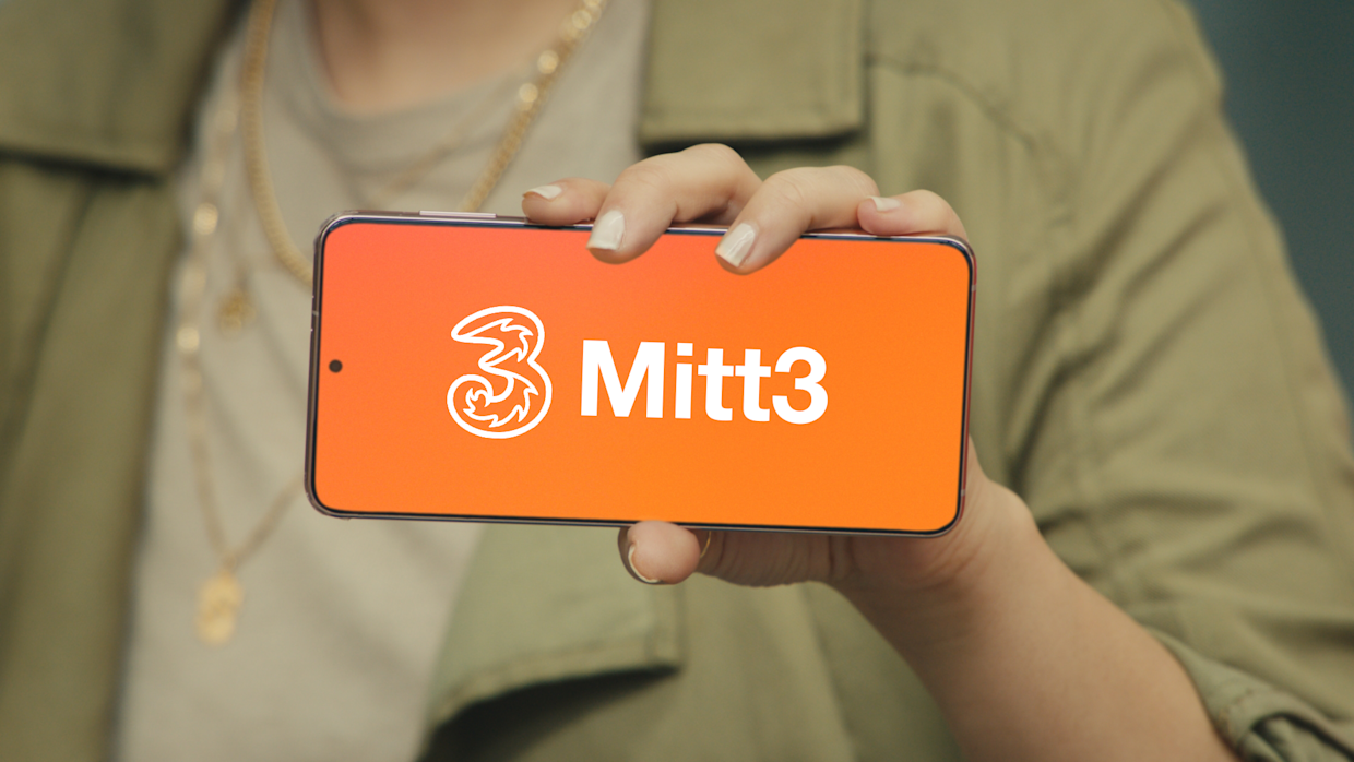 En person håller upp sin svarta smartphone med Tre Företagsloggan och Mitt3 i vitt med orange bakgrund.