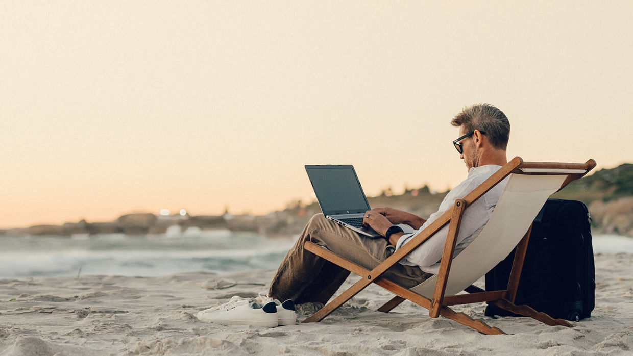 Man sitter på stranden i en solstol och jobbar med sin dator.