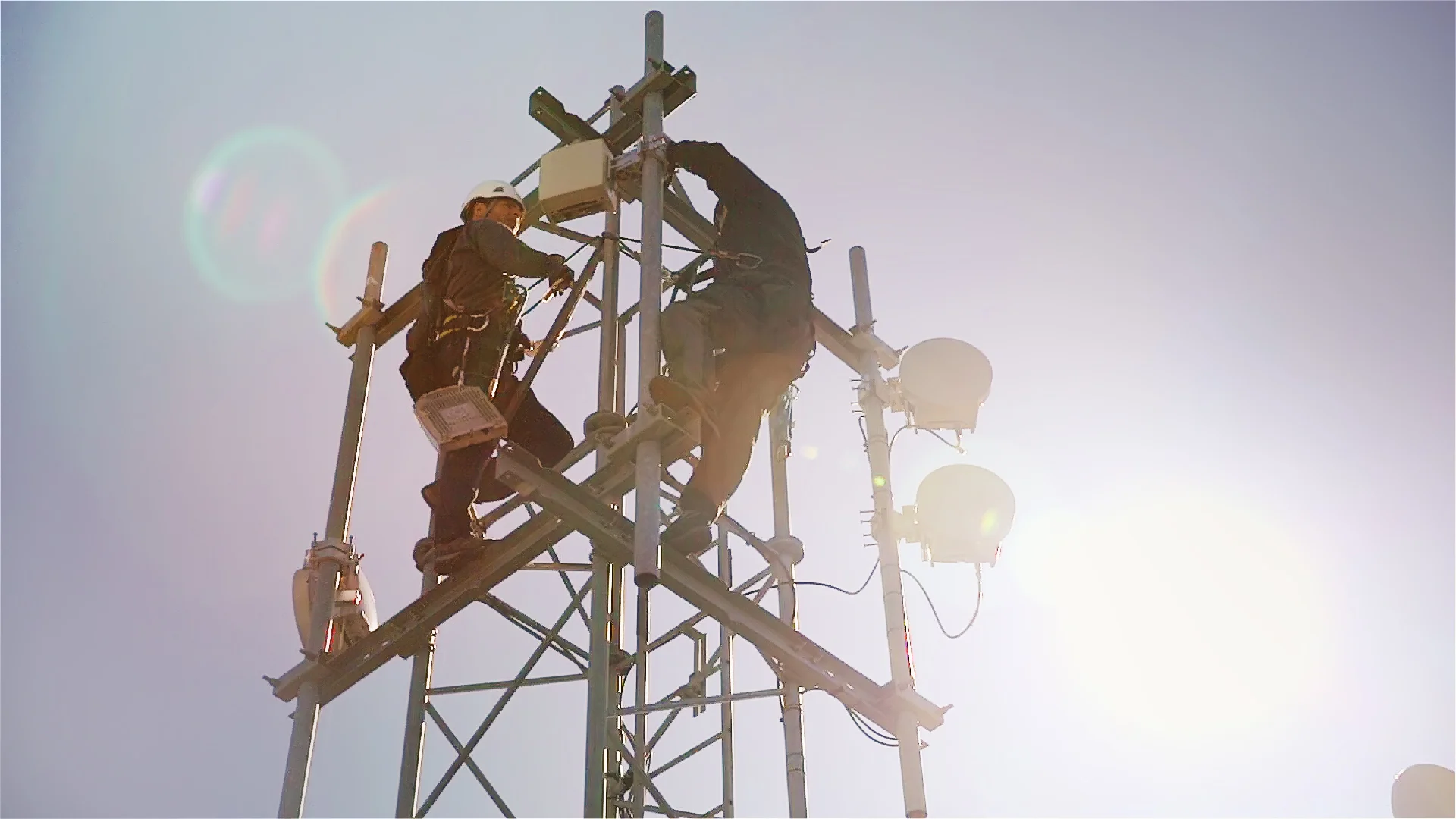 Två arbetare i säkerhetsutrustning justerar utrustning högst upp i en telefonmast. Himlen är klar och solen skapar ljusfläckar i bakgrunden.