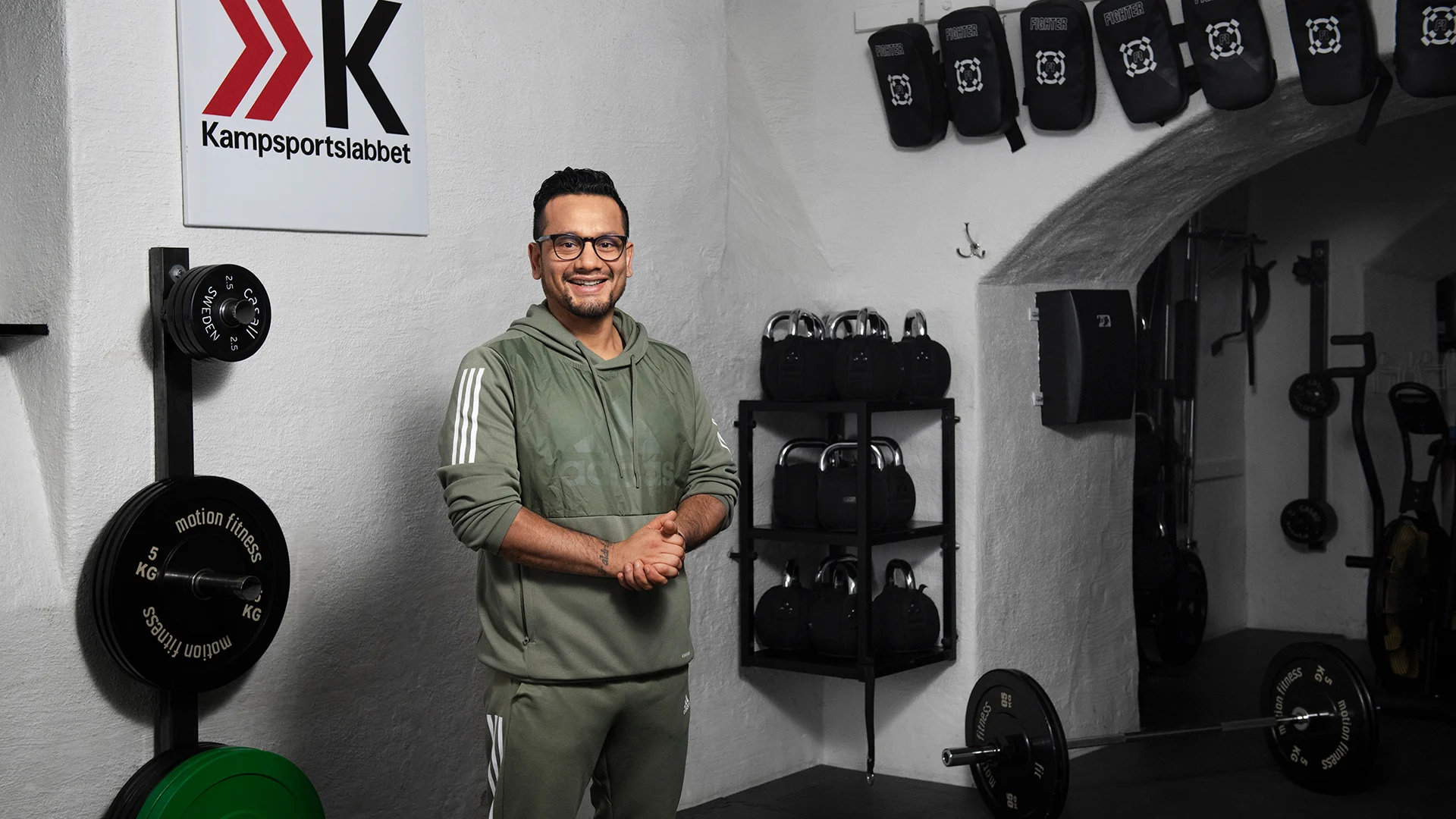 Man i mossgröna träningskläder och glasögon i en träningslokal. Olika typer av träningsredskap hänger på väggarna eller står på golvet.