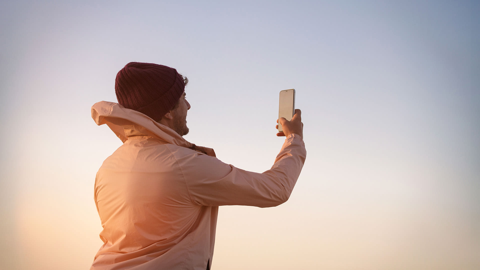 Man i svart mössa och vit vindjacka håller i en mobil. Det ser ut att vara soluppgång/solnedgång och som att han fotar den.