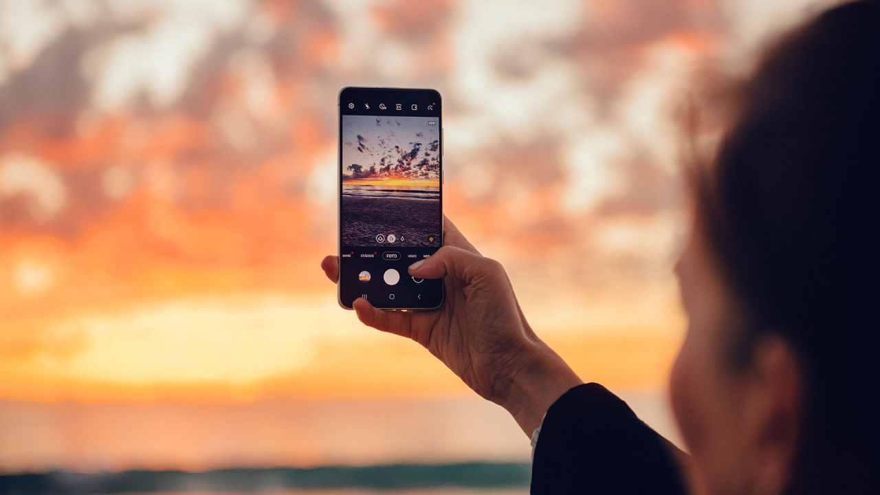En kvinna håller en mobil i handen och fotar en solnedgång.