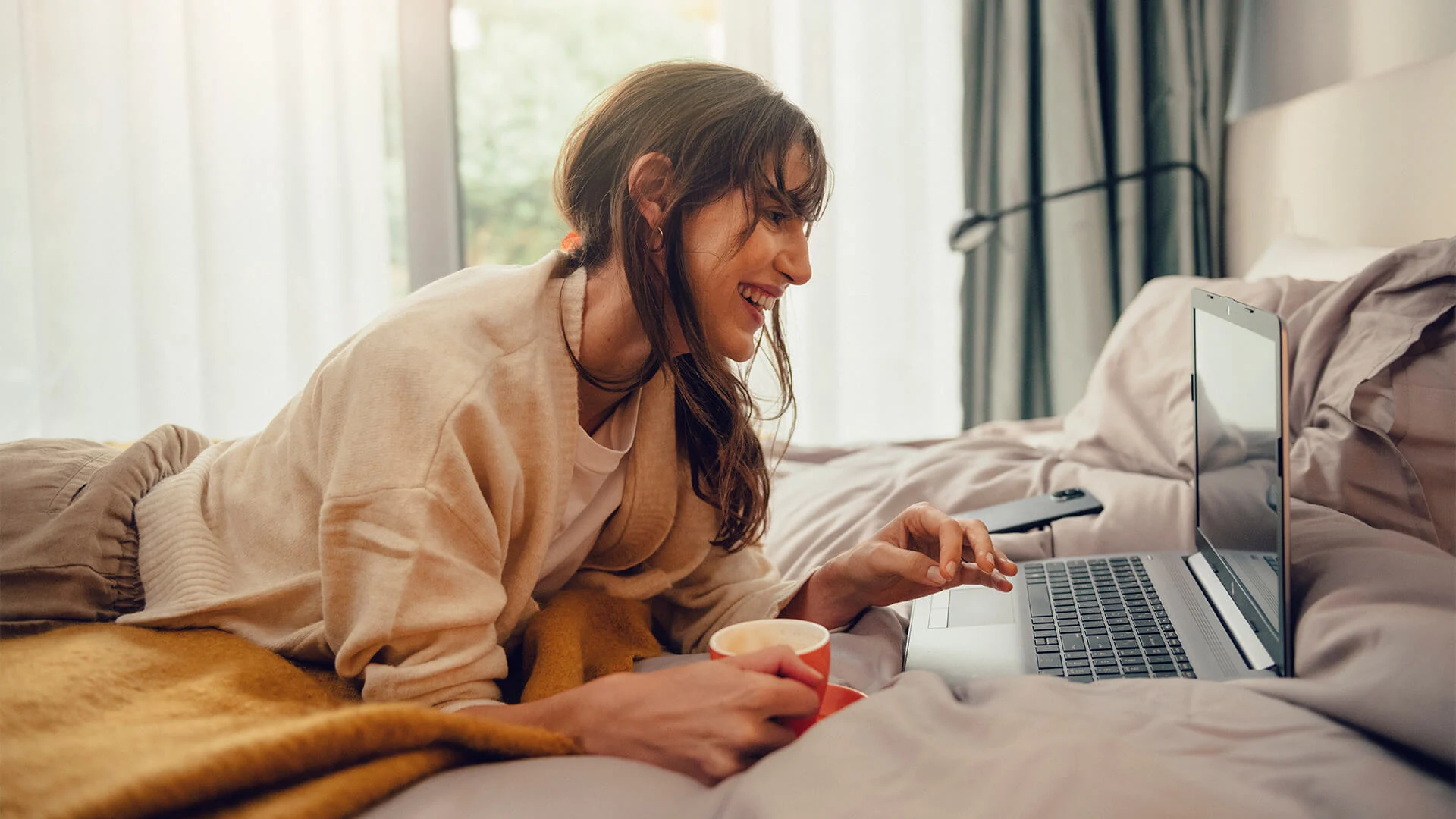 Kvinna ligger på en säng med en kaffekopp i handen och tittar på en dator.
