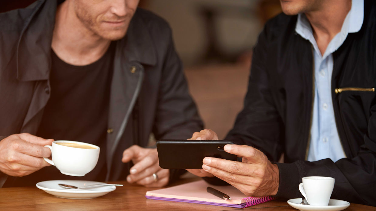 Två personer i mörka kläder sittandes vid ett bord med varsin kaffekop. En av personerna visar den andra något på en mobiltelefon.