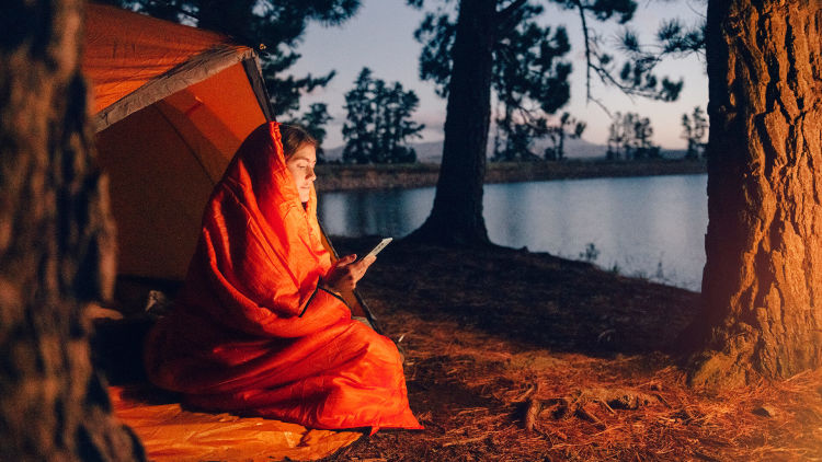 En kvinna sitter framför ett tält vid vattnet med en sovsäck runt sig. I handen håller hon en mobil som hon tittar på.