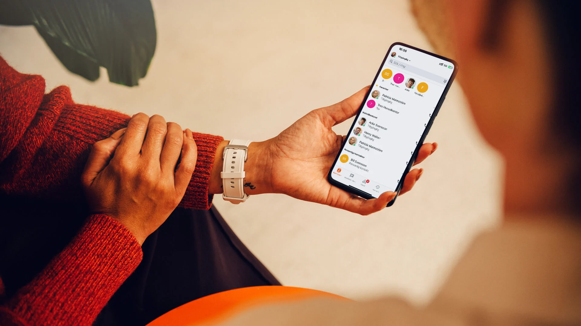 Person håller en smartphone med en appen 3Koppla öppen, klädd i röd tröja och vit klocka, sittande inomhus.