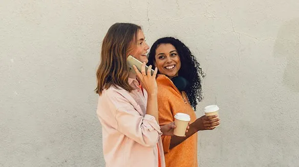 Två tjejer går med en latte i handen och ena pratar i telefon.