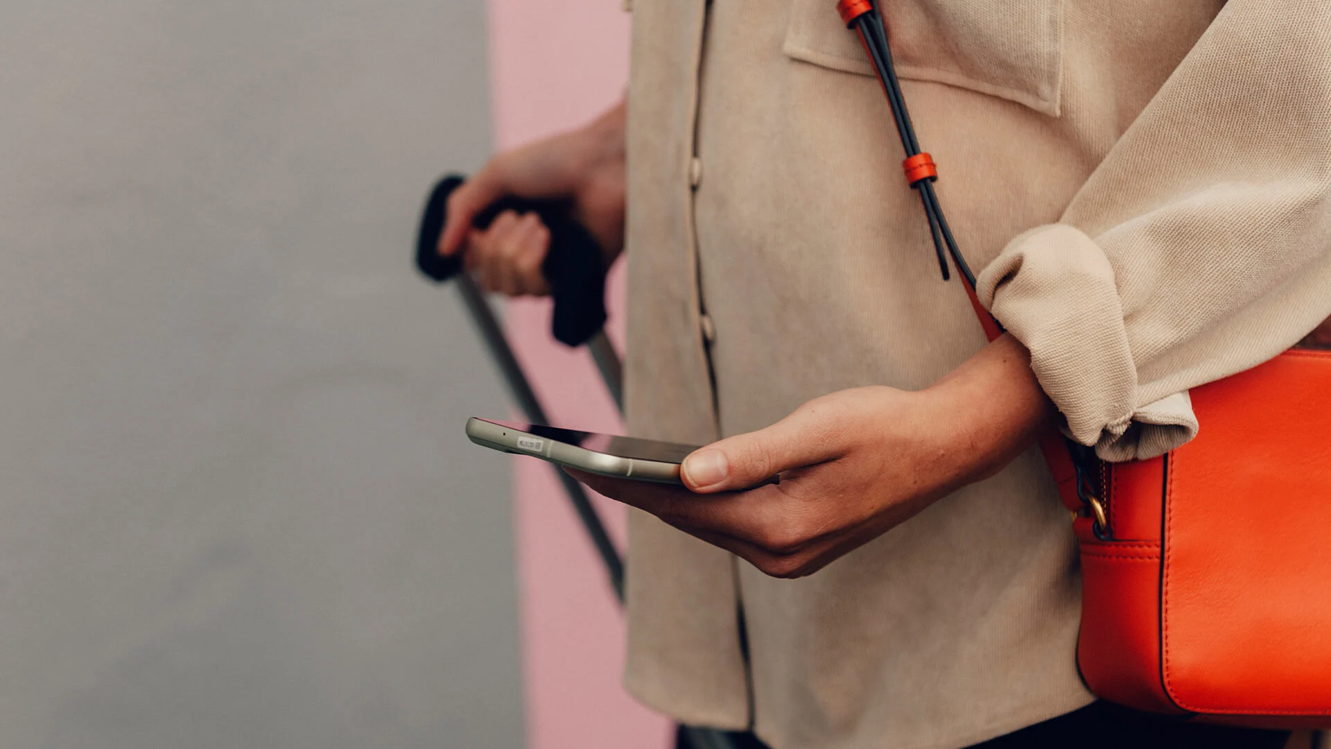 Person i beige skjortjacka håller en mobil i ena handen och drar en resväska i den andra. En orange väska hänger över axeln.