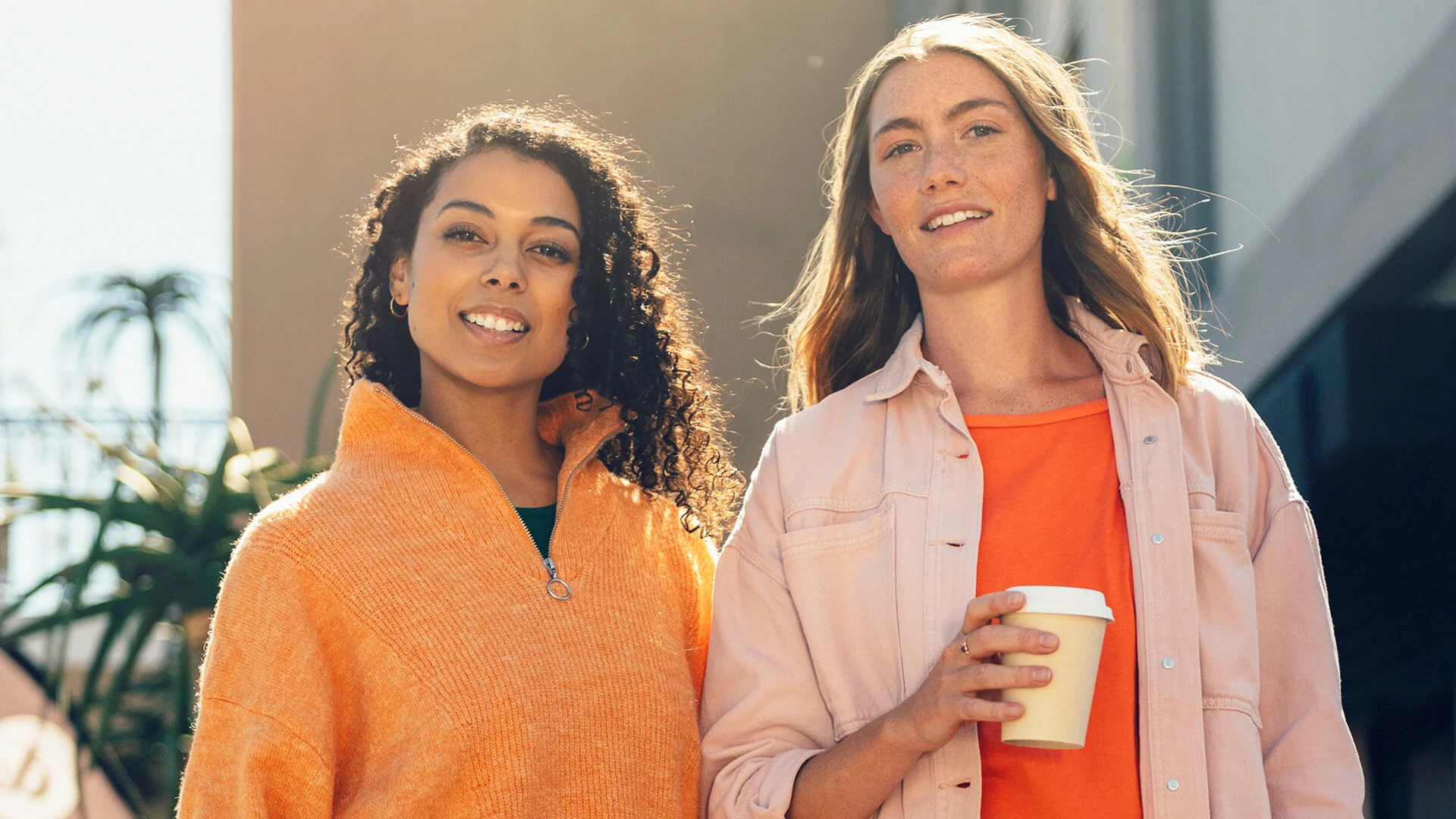Två kvinnor tittar in i kameran och ler, den ena håller i en take away-kaffe.