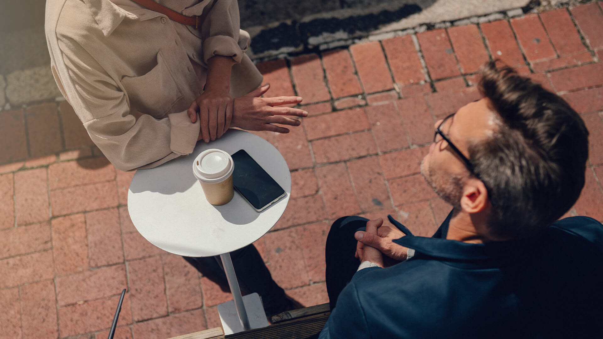 Två personer samtalar utomhus över en kaffe, en mobil ligger på ett vitt bord.
