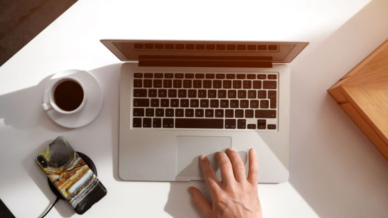 Ett skrivbord med en dator, en mobil som ligger och laddar på en trådlös laddplata och en kopp kaffe med fat.