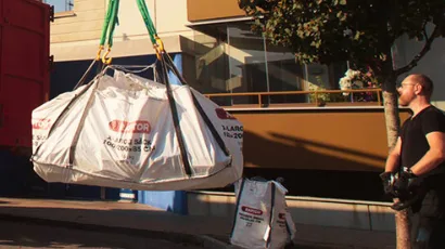 En man står bredvid en stor vit säck som lyfts med kran framför en byggnad. Säckarna har text och logotyp.