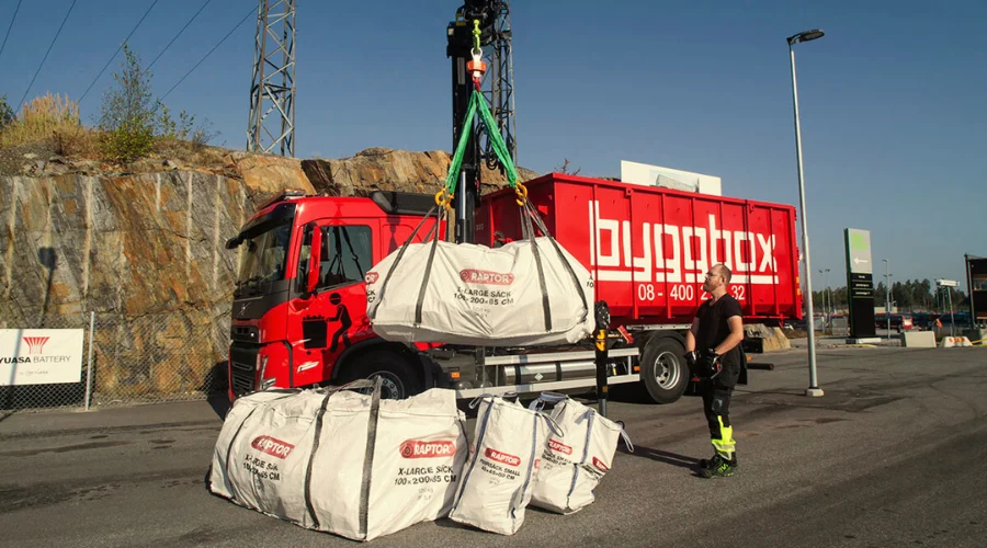 En man står bredvid en stor vit säck som lyfts med kran framför en byggnad. Säckarna har text och logotyp.